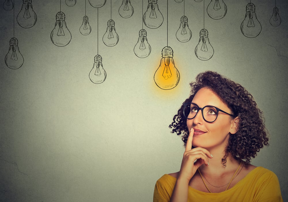 Thinking woman in glasses looking up with light idea bulb above head isolated on gray wall background Thinking woman in glasses looking up with light idea bulb above head isolated on gray wall background